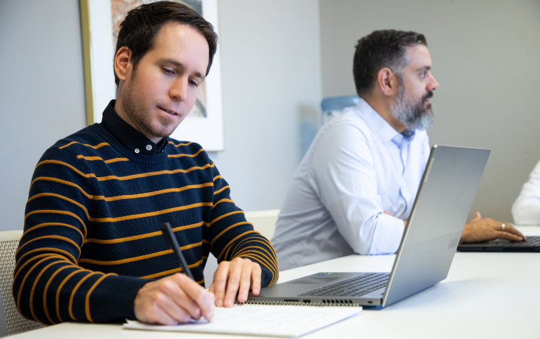 Two men attending a meeting, and the one closer is taking notes
